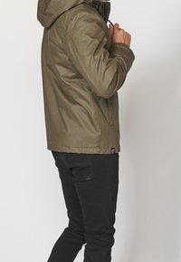 Mężczyzna w oliwkowo-zielonej kurtce z kapturem i czarnych spodniach, poprawiający kołnierz kurtki, stojący na jednolitym jasnym tle.