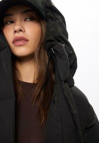 Giacca a vento nera con cappuccio alto e arricciato, dotato di cordoncino, indossata sopra una maglia scura. Texture liscia e design minimale.