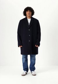 Tumši zils vilnas pārsegu mētelis ar piegrieztu siluetu, dubulto pogu aizdari un sānu kabatām, valkāts pār baltu T-kreklu un zilām džinsām.