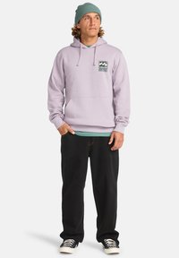 Felpa lavanda con tasca frontale e patch del logo, abbinata a pantaloni neri e una maglietta teal sotto. Un berretto verde completa il look.