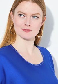 Top blu, a maniche corte con scollatura arrotondata e dettagli di cucitura sottili. Orecchini a cerchio dorati con un design liscio e curvo.