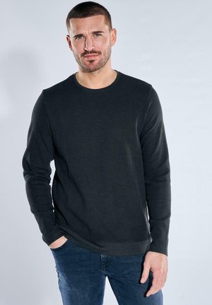 Man with short hair and beard wearing a black long-sleeve ribbed shirt and blue jeans, standing with one hand in pocket.