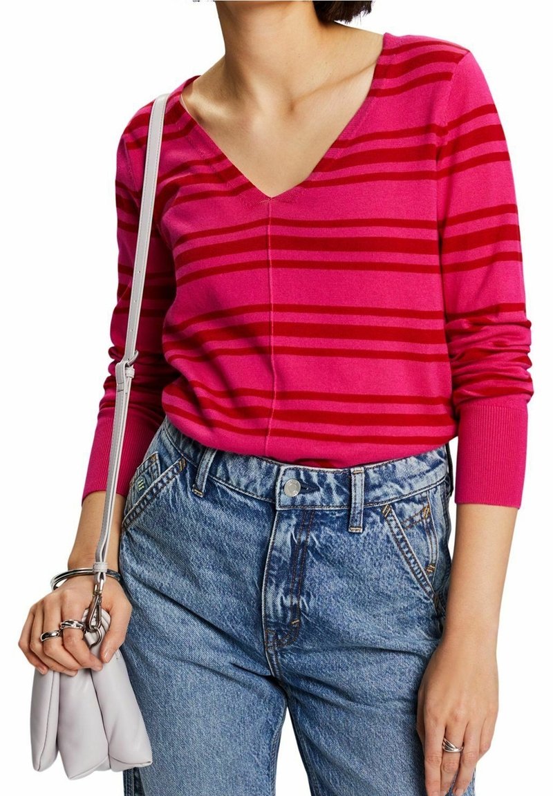 Rosa gestreifter Pullover mit V-Ausschnitt und langen Ärmeln, kombiniert mit hoch taillierten blauen Jeans aus Denim. Weiße kleine Handtasche und silberne Ringe sichtbar.