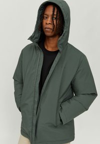 Green insulated jacket with hood, featuring a front zipper, side pockets, and a smooth texture. Worn over a black shirt.