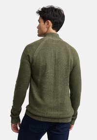 Olivengroene gebreide trui met een hoge kraag, raglanmouwen en geribbelde boorden en zoom. Gestructureerde stof met subtiele glansdetails.