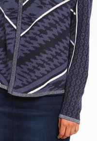 Stickad tröja i mörkblått med ljusgrå detaljer. Har geometriska mönster; texturerad tyg med långa ärmar och knäppning framtill.