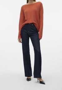 Femme portant un pull long à manches longues orange, un jean large noir et des escarpins noirs à bout pointu, se tenant contre un fond blanc.