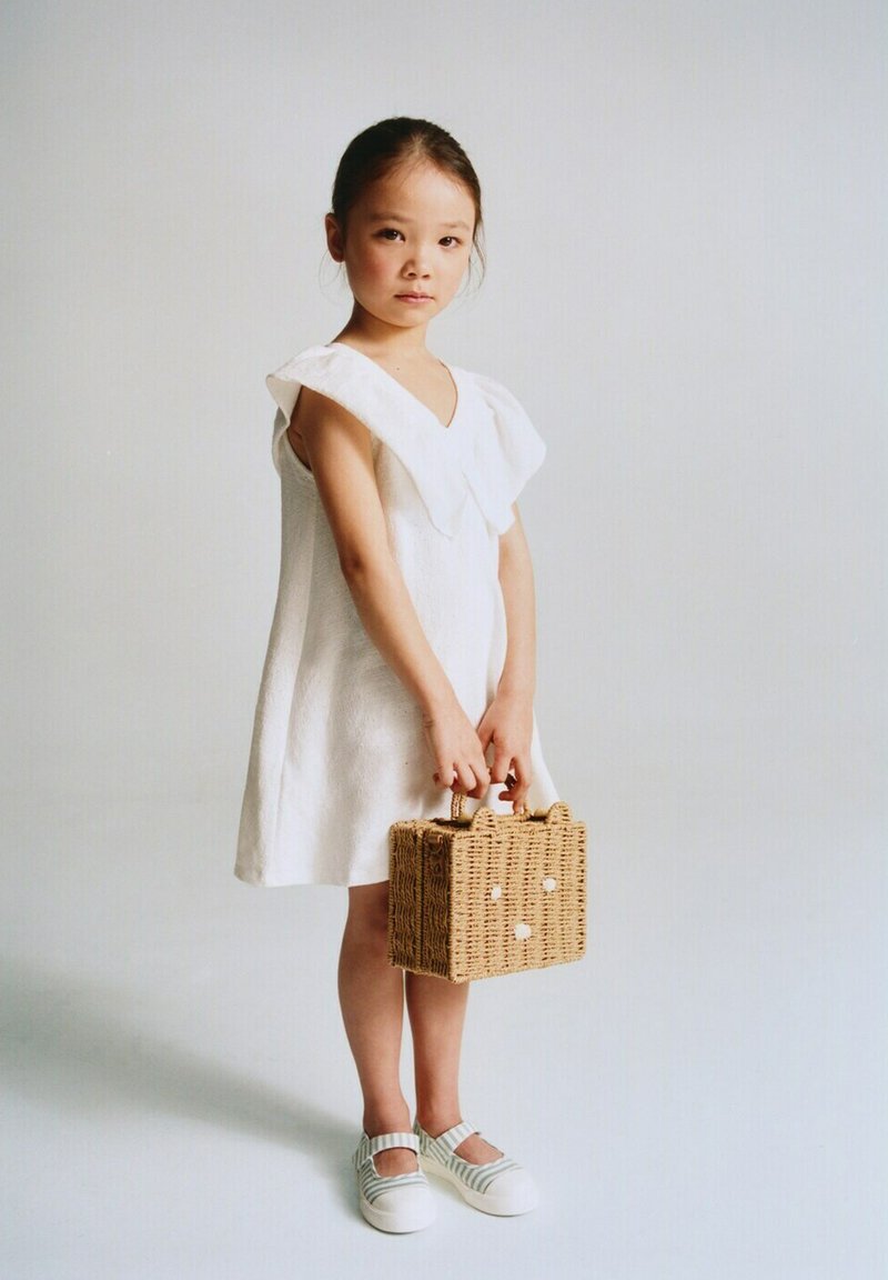 Jeune fille en robe blanche et chaussures rayées tenant un sac à main rectangulaire tissé, debout devant un fond clair uni.