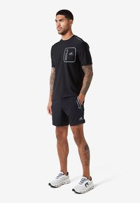 Muž s tetovaniami stojí v čiernych športových šortkách a tričku s detailom vrecka, doplnený bielymi športovými topánkami.