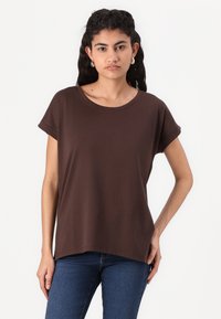 Brown short-sleeve t-shirt with a round neckline, loose fit, and textured fabric. Worn with blue jeans, featuring rolled sleeves and a straight hem.