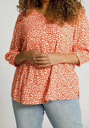 Frau trägt locker sitzende orange-weiße gepunktete Bluse mit Dreiviertelärmeln, kombiniert mit hellblauen Jeans, Hände verschränkt.