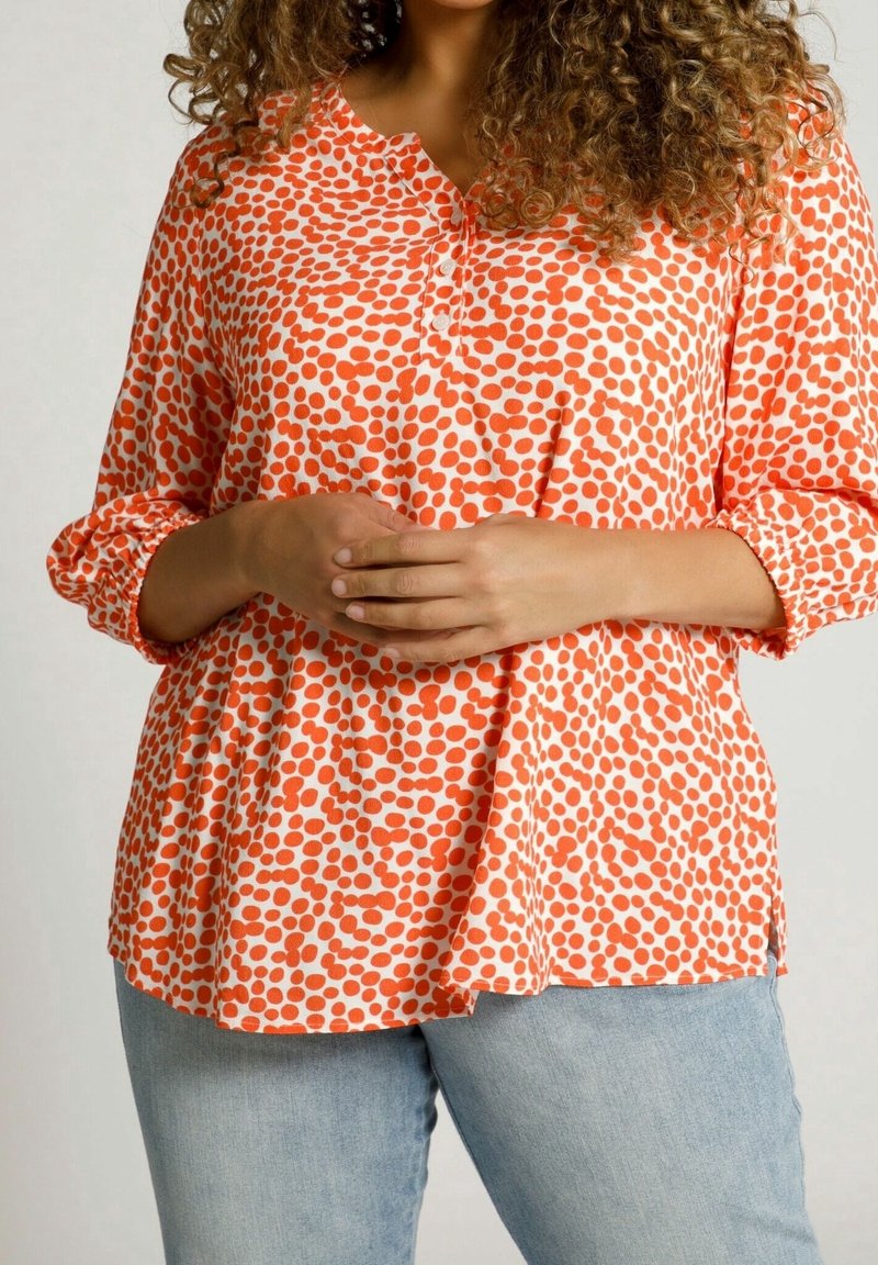 Femme portant un chemisier ample orange à pois blancs avec manches trois-quarts, associé à un jean bleu clair, mains jointes.