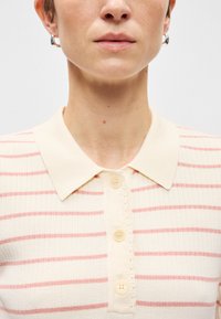 Πλεκτή πόλο μπλούζα σε κρέμα χρώμα με κόκκινες ρίγες, διαθέτει γιακά, επτά κουμπιά και υφασμάτινη υφή με οριζόντιο σχέδιο ραβδώσεων.