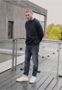 Černá mikina s malým logem na hrudi, modré džíny a bílé boty se zelenými akcenty, stojící na dřevěné střešní terase.