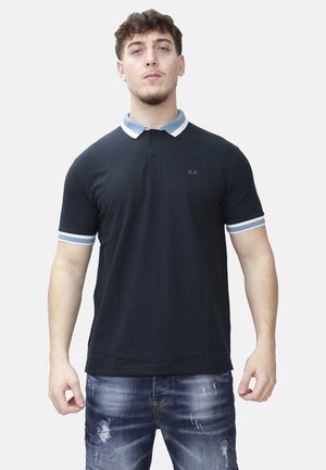 Man draagt een zwarte polo met een kraag en mouwen met blauwe en witte strepen, en versleten blauwe spijkerbroek, staand tegen een effen achtergrond.