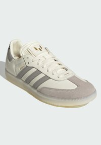 Zapatilla blanca y gris con materiales de cuero liso y gamuza, que presenta tres rayas grises, una puntera texturizada y un logo dorado en la lengüeta.