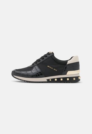 Baskets noires avec des accents beige au talon et sur la semelle, des clous dorés sur la semelle, des panneaux latéraux texturés, des lacets et le logo "Michael Kors" sur le côté.