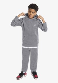 Gray hoodie with kangaroo pocket, front logo, matching gray sweatpants, and black/red sneakers. Model adjusting hood.