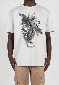 Camiseta de color gris claro con un gráfico en blanco y negro de un ángel rodeado de texto en espiral y patrones similares a nubes. Material de algodón.