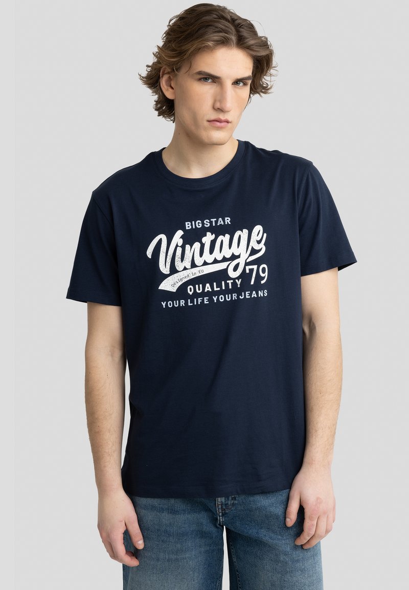 Tamsiai mėlynas medvilnės marškinėliai su trumpomis rankovėmis, ant kurių priekio yra balta vintage stiliaus grafika ir klasikinis apvalus iškirpimas.