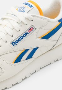Białe skórzane sneakersy z niebieskimi i pomarańczowymi akcentami, z teksturowaną powierzchnią, wyraźnym logo i klasycznym sportowym designem.
