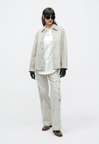 Hellgraue Wildlederjacke über einem weißen Hemd, kombiniert mit hellgrauen Cargohosen und schwarzen Handschuhen. Accessoires umfassen dunkle Sonnenbrillen.