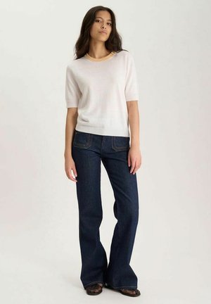 Femme aux longs cheveux foncés portant un haut léger à manches courtes, un jean évasé foncé et des chaussures noires, debout devant un fond uni.