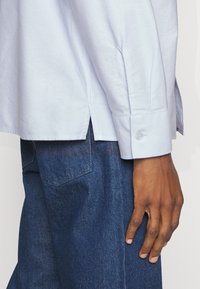 Camicia azzurra chiaro con maniche lunghe, spacchi laterali e polsini con un solo bottone. Indossata sopra jeans in denim blu scuro con dettagli sulle tasche.