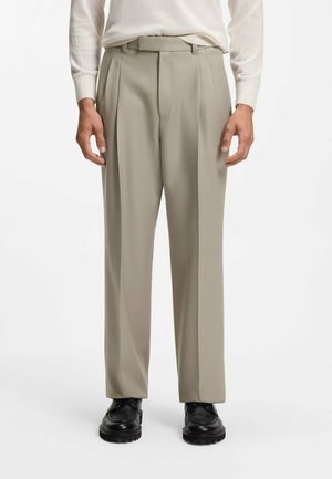 Hombre con pantalones de vestir plisados beige, zapatos negros y camisa blanca de manga larga, de pie frente a un fondo blanco liso.