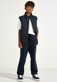 Marineblå puffer vest over en hvid T-shirt, parret med mørke sweatpants og blå sneakers med hvide og gule accenter.