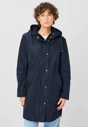 Frau trägt eine marineblaue Kapuzen-Regenjacke mit Druckknöpfen und Seitentaschen, kombiniert mit hellblauen Jeans, steht vor einem schlichten Hintergrund.