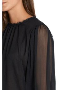 Blouse noire avec un col transparent froncé et des manches longues légèrement bouffantes. Le tissu présente un motif texturé à rayures verticales.
