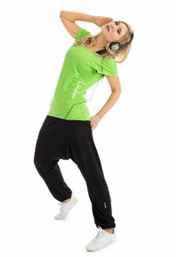 Groene top met korte mouwen en witte tekst, gecombineerd met zwarte baggy broek. Draagt grijze koptelefoon en witte sneakers, dynamisch geposeerd tegen een witte achtergrond.