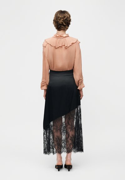 Άτομο που φοράει μια ρουφηχτή μπλούζα ροδακινί με μακριά μανίκια και μια ασύμμετρη μαύρη φούστα με διαφανή δαντελένια λεπτομέρεια, στέκεται με την πλάτη προς τα εμπρός.