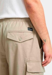 Pantalones cortos de cargo beige con una cinturilla elástica, que presentan un bolsillo trasero con cierre de botón y un parche con logo. La tela parece suave y texturizada.