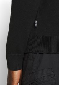 Jersey de lana negro con puños y dobladillo acanalados, que presenta una pequeña etiqueta lateral con el nombre de la marca. La tela parece suave y con textura.