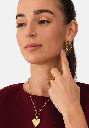 Frau mit dunklen Haaren trägt goldene, herzförmige Ohrringe, eine passende Herzanhänger-Halskette und mehrere Goldringe an ihrem Finger.