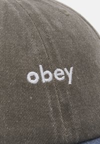 Khaki színű pamut sapka ívelt szélekkel, elöl fehér hímzett "obey" felirattal. A textúrája sima, látható varrási részletekkel.