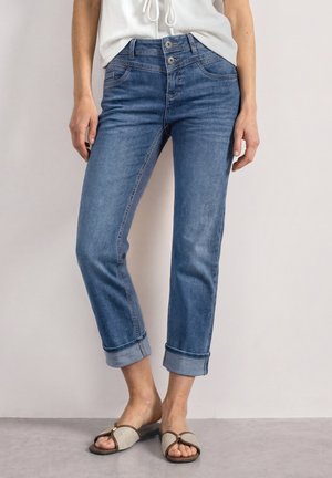 Femme portant un jean bleu retroussé avec une taille à double bouton et des sandales beige ouvertes à l'avant, debout devant un fond clair.