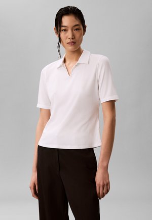 Kobieta o ciemnych włosach ubrana w białą koszulę z krótkim rękawem i kołnierzykiem oraz czarne spodnie, stoi na tle jednolitego szarego tła.