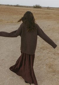 Ruda vilnos megztinis su plačiomis rankovėmis, derinamas su laisvai krentančia vyno spalvos sijonu. Audinys atrodo minkštas ir tekstūruotas prieš negyvenamą peizažą.