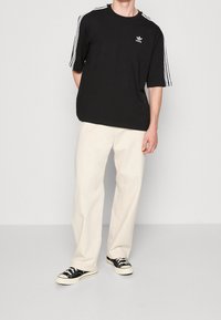 Svart kortärmad t-shirt med vitt tre-stripersdesign längs axlarna och liten logotyp på bröstet, matchad med ljusa byxor och svarta sneakers.