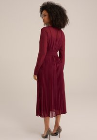 Long burgundy pleated dress with long sleeves, fitted waist, and sheer fabric. Worn with gray high heels, showing a smooth texture.