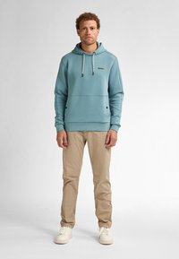 Lichtblauwe hoodie met een voorzak, trekkoord capuchon en zichtbaar logo. Gecombineerd met beige broek en off-white sneakers.