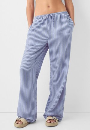 Kvinde iført løstsiddende blå og hvide bukser med vertikale striber, snor i taljen og beige sandaler, stående med hænderne i lommerne.