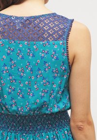 Robe sans manches avec un imprimé floral turquoise, superposition en dentelle au dos en bleu marine, ceinture froncée et bordure à pompons le long des manches.