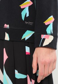 Zwarte trui met kleurrijke bladpatronen in roze, blauw en groen; voorzien van gestikte details en logo op de zoom; gedragen met een zwarte geplooide rok.