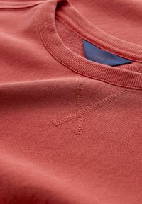 T-shirt in cotone corallo con collo rotondo e cuciture visibili. Include un'etichetta blu sul collo e dettagli di texture nel tessuto.