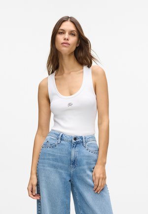 White tank top with wide straps and a scoop neckline, featuring a small embroidered logo. Paired with light blue high-waisted jeans, embellished with studs.