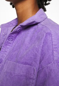 Purple corduroy shirt with a collar, front button closure, and a chest pocket, featuring vertical ridges for texture.